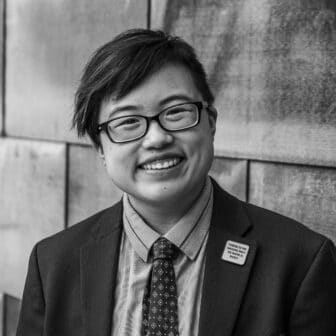 Black and white photo of Lydia X. Z. Brown. Lydia smiles and tilts their head slightly to the side, looking confidently at the camera. They are a young-ish East Asian person with a streak of teal in their short black hair, wearing glasses, a cobalt blue jacket and navy tie, with a blue copper wall behind them. Photo by Sarah Tundermann.