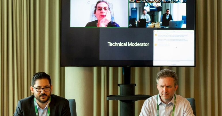 A hybrid session at RightsCon with two people in a room and a TV monitor with other people participating